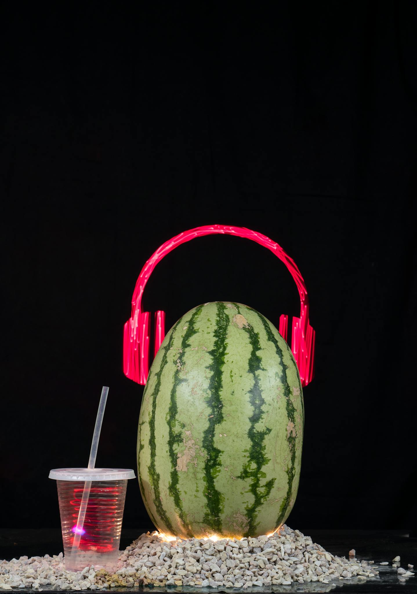 Creative watermelon with red headphones and drink against black backdrop.